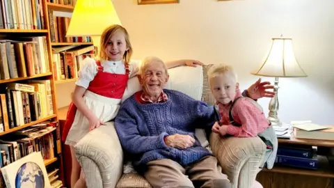 Slater family An elderly man on a chair, with two blonde children perched on either arm. They are smiling, with bookshelves in the background. 