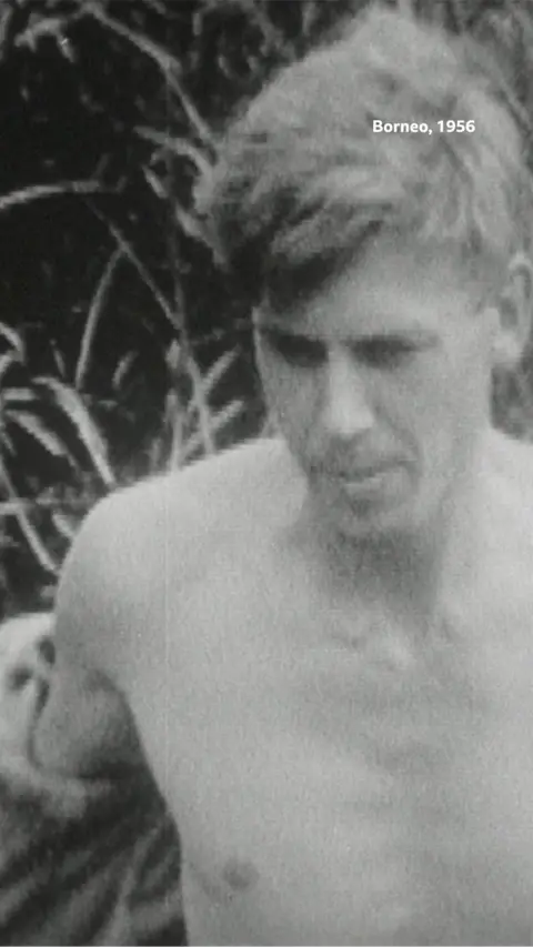 A close-up of Sir David Attenborough topless walking through a forest