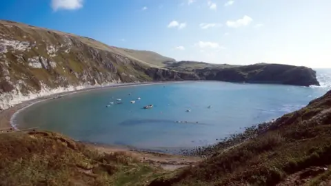 View across Lulworth Cove from the nearby cliffs. The cove is almost circular and surrounded by grass-covered cliffs. To the right of the picture is a gap where the water flows out to the open sea. In the centre are about 10 small boats.