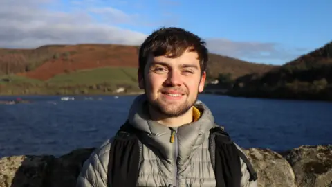 A young man wearing a grey jacket and black rucksack stood in front of a lake and a hill