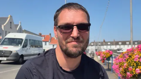 Jason Horsepool is outside. He is wearing shaded reading glasses and a navy blue T-shirt, stood outside next to harbour with colourful yellow and pink flowers in sunny conditions.