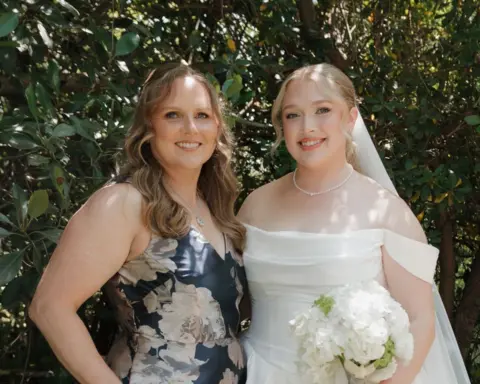 Kylee Lynn Photography Hannah and her mother at Hannah's wedding. Her mother is wearing a blue dress with white flowers. Hannah on the right is dressed in a white wedding dress holding a bouquet