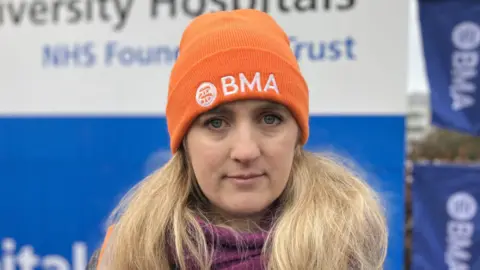 Dr Hanna Copley is unsmiling and looking directly at the camera. She is wearing an orange hat that says BMA on it and is stood in front of a sign. 