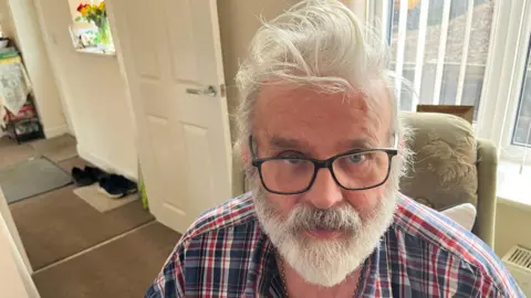 John Devine/BBC Michael Weston has wavy grey hair and matching beard and moustache, he has black framed glasses on and a tartan shirt with a gold chain around his neck, he is in a sitting room, with a door open and shoes visible on a mat.