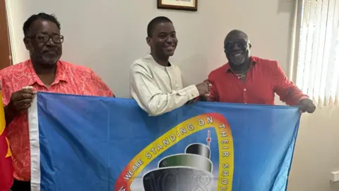 Charles Dacres Nigel Guy (left) and Charles Dacres (right) holding the flag with St Kitts and Nevis PM, Dr Terrance Drew (centre)  