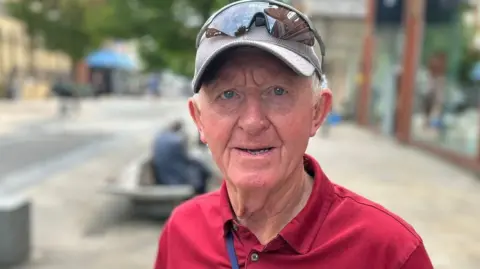 Colin Richardson, with white hair, wears a grey baseball cap with sunglasses on top. He is sporting a red open-necked shirt.