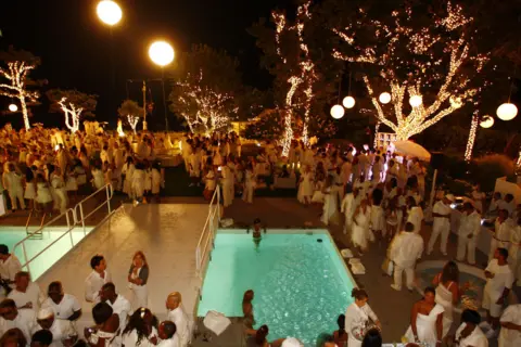 Getty Images A White Party at the Hamptons in New York state, seen from a high vantage point, showing a large crowd all dressed in white, standing around a large swimming pool, which has a white bridge constructed over it. The tree trunks and branches are wrapped in fairy lights with large globe pendant lights hanging from some of them.