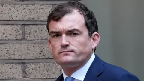 Getty Images A man looks at the camera as he walks past a brick building. He has short dark hair and is wearing a blue suit with a white shirt and blue tie. He is frowning.