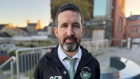 This is a blurred in focus photo. The background is buildings that are sun lit and the sky is blue. Emett stands in front of railings. He is wearing a blue tie and white shirt with a black raincoat with a green and white badge on the raincoat. 