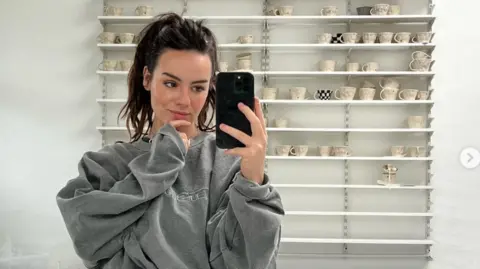 Mollie Warrington A young woman with dark hair, pulled back into a high ponytail, wearing a grey sweatshirt takes a mirror selfie, behind her is shelving filled with handmade neutral mugs.