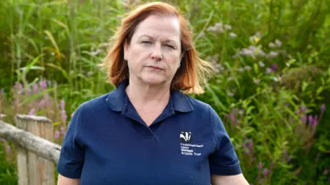 BBC Natalie Buttriss is wearing a navy polo top and is standing in front of a bank of high reeds, staring straight at the camera.