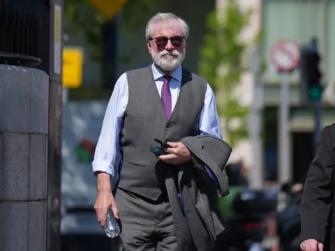 PA Media Gerry Adams - a man with grey hair, grey beard - wearing dark sunglasses and a grey suit with matching trousers and waistcoat, a blue shirt and purple tie. He is holding his jacket across his arm, it is sunny, and he has a bottle of water. Background is out of focus but it is outside a courthouse.