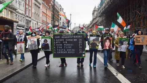 Getty Images Protesters walk in a row down a city street holding pictures of six of the prisoners on hunger strike, with a large black placard at the centre listing their names. 