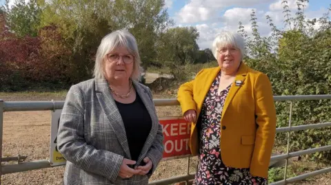 Liberal Democrats Two women face the camera. They stand in front of a gate that says "keep out" There are a number of trees in the background.