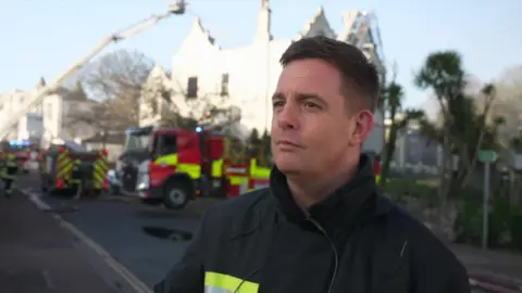 Ben Williams is looking to the left with the hotel behind him. There is a firefighter on an aerial ladder behind him. 