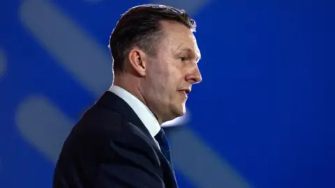 Russell Findlay, who has short black hair, in profile against a blue background. He is wearing a business suit