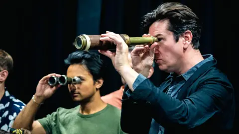  Lian Furness  Ed Hawkins looking through a telescope during a rehearsal. He is wearing a blue shirt and a dark blazer. Other cast members can be seen in the background, including one looking through a pair of binoculars