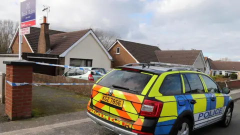 Pacemaker A row of residential homes with a yellow, blue and orange police car on the kerb outside. there is blue and white police tape and a 'sold' house sign. The houses are small bungalows with red bricks, brown roofs and cream paint.