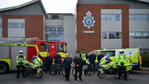 A group of police and emergency services together with electric bikes stood in front of a police van and a fire engine
