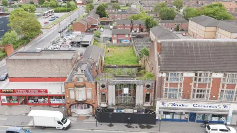 An aerial view of the theatre. At the front is the bomb-damaged facade with scaffolding. Construction workers are standing on wasteland behind, which is the space where the auditorium once stood. To the left of the theatre is a pub and a food store and to the right is a ceramics shop.