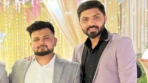 Nirav Patel Two men standing together wearing suits. Nirav is wearing a grey suit and Vaibhav wearing a purple suit.