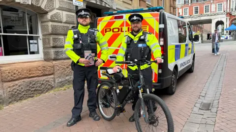 George King/BBC PC Lewis Finch and PC Matthew Dilks, who are both wearing a police uniform, standing in front of a police van and behind an e-bike that they seized moments before.