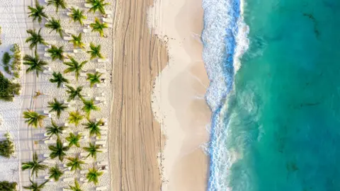 A beach in Cancun. The picture shows white sand, palm trees and an aqua sea with waves breaking on the shore