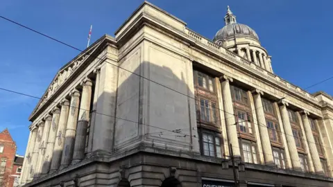 An image of Nottingham's council house in the city centre from the front corner of the building showing columns at the front and sides of the building with a dome and clock visible in the top right-hand side of the frame. It is a sunny day with no clouds and a Union Jack flag can be seen at the top of the building's frontage. 