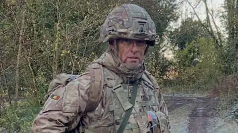 Scott Caswell Scott Caswell stands on a dirt track in a wooded area in full military camouflage uniform, wearing a camouflage rucksack and helmet. 