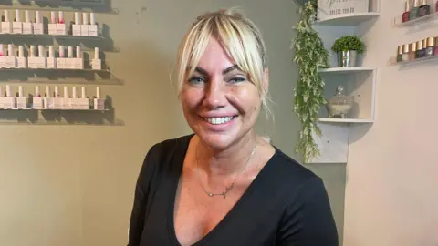 A woman with blonde hair tied up in a bun smiles at the camera inside a nail salon. She is wearing a black V-neck top and a delicate necklace. Behind her, shelves display rows of nail polish bottles and a sign reads 'sparkle'.
