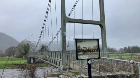 BBC Gower Bridge is seen with a sign explaining its historical significance. In the background there are fields and trees on a foggy day in Llanrwst.