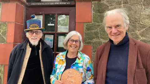 BBC Two men and one woman stood in front of a red brick framed door. One man is wearing a blue top hat with the word "Dennis" on it and the other has white hair and a brown jacket. The woman in the middle is wearing a multi-coloured jacket and is holding an oversized copper coin