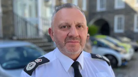 BBC Damian Kitchen - A man with grey and black hair looking at the camera. He has stubbly beard and is wearing a police uniform with silver insignias on black lapel badges. He is wearing a black tie on his white shirt. 