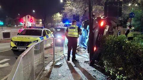Cambridgeshire Police A police car on the road with a car on its side on the roundabout - inside the metal barriers. Two police officers can be seen on the scene.