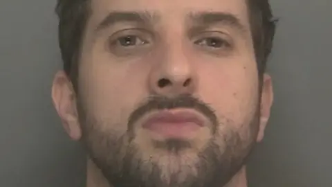 Merseyside police Thomas' mugshot after arrest. He stands before the camera against a grey background. He has dark facial hair and hair.