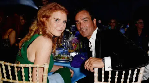 Getty Images Sarah Ferguson and Manuel Fernandez sit at a table laden with glasses, plates and a blue table cloth. She wears a green dress, he wears a black velvet jacket and a white shirt open at the neck. They sit at wickerwork-style chairs, and are looking over their shoulders at the camera