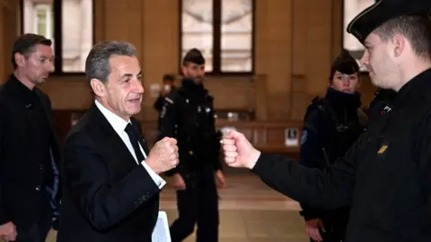 Getty Images Nicolas Sarkozy fist bumps a policeman as he arrives at court for his case