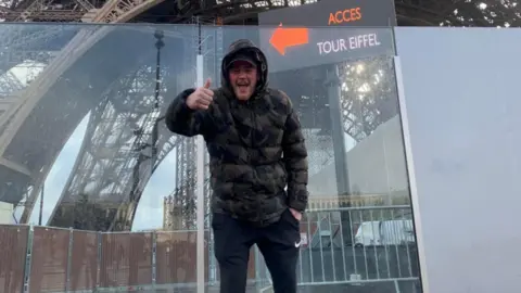 Megan Holmes Connor Walker, with his thumbs up in front of the Eiffel Tower.