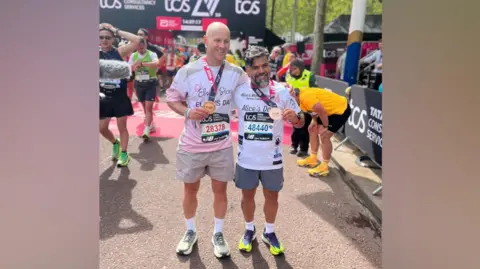 BBC David Stancombe and Sergio Aguiar pose with their arms around each other as they hold their medals at the finish line. Other runners mill around behind.
