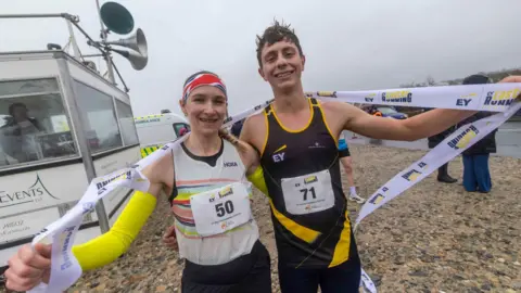 A picture of Louise Small (left) and Luke Holmes (right) celebrating after their record breaking runs. Louise is wearing a white running shirt and wearing a headband. Luke is wearing a black and yellow shirt. They are both holding a ribbon and are stood next to one another on a rock like surface.
