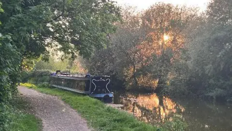 Ruth A narrowboat moored on a bend in a river lined by trees. The low orange sun is shining through the trees.