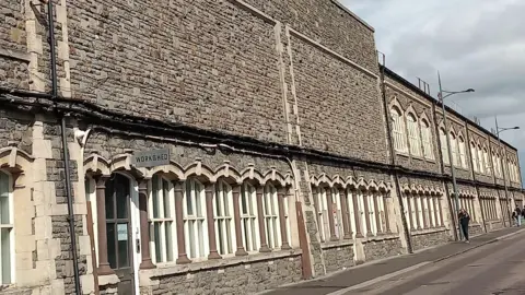 Jack Hayward An image of a tall brown and grey stone wall, the bottom section of which includes a row of arched windows and a brown door. A sign over the door reads "workshed".