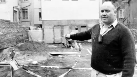 Getty Images The decomposed body of a young woman was found by workers in the back yard of a house in Fitzhamon Embankment, Cardiff. The body was found in the corner where police have erected a polythene tent. Unfortunately, building activity has ruined much of the evidence. Pictured is Paul Bodenham of Roath Housing building contractors, who found the body, showing the corner of the yard where it was found. It is a black and white image of him pointing to where he found the body (in the garden)