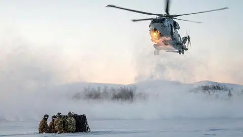 A helicopter is flying over a snowy landscape. Some marines are sitting together on the snow.