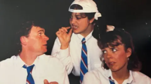 Sheringham Theatre Jones (centre) wore a back-to-front white baseball cap, a white school shirt and striped school tie. She is looking directly at the male actor, also dressed in the same uniform, in front of her and has her hand in front of her face. A female actor, also in the uniform, pulls a quizzical-looking face.