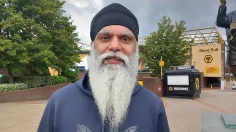 BBC Manny Singh Kang standing in front of the Wolves football stadium. He has a dark head covering, wears a blue top and has a long white beard