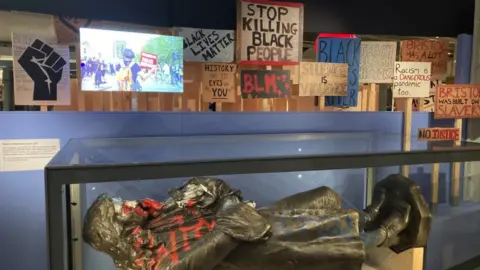 BBC Image of the exhibition. The Colston statue is in a glass case and protest signs can be seen behind it.