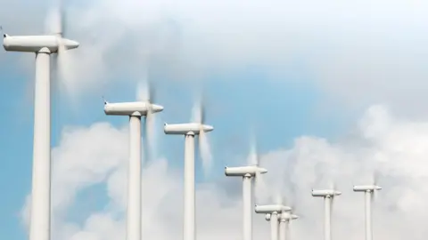 XXLPhoto/Getty Creative A row of white wind turbines rotating, pictured in front of a blue sky with fluffy white clouds. 