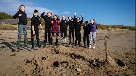 Dru Dodd Eight children jumping in the air on a beach. They are standing in front of a piece of beach art they have created. It is a sunny day and two sheds are visible behind them. 