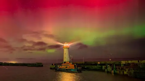 BBC weather watcher / Ray McDonald The Northern Lights lighting up the sky in pink and green colours. A white lighthouse and a harbour is at the bottom of the image. 